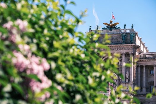 View of the ornate facade of the Hofburg Palace with its golden eagle statue framed by blooming pink flowers and green leaves under a clear blue sky in Vienna Vienna Austria. View of the ornate facade of the Hofburg Palace with its golden eagle statue framed by blooming pink flowers and green leaves under a clear blue sky in Vienna Vienna Austria.