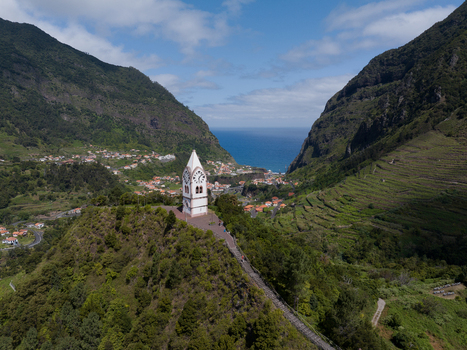 Aerial view of the Capela de Nossa Senhora de Fatima clock tower perched on a green hill overlooking a coastal village terraced hillsides and the Atlantic Ocean in Madeira. Aerial view of the Capela de Nossa Senhora de Fatima clock tower perched on a green hill overlooking a coastal village terraced hillsides and the Atlantic Ocean in Madeira.
