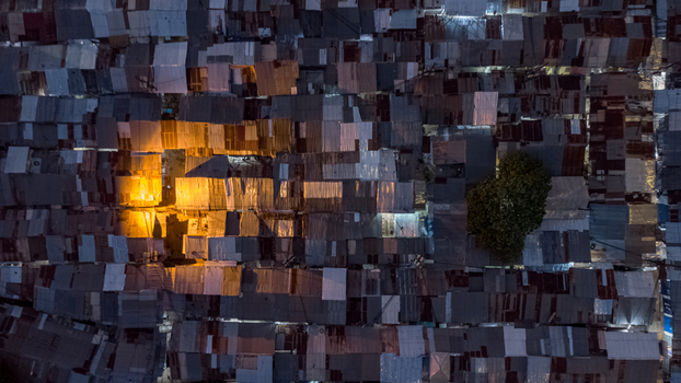Aerial view of a dense urban neighborhood with corrugated metal rooftops and a single tree illuminated by warm artificial light at night in Tamale Northern Region Ghana. Aerial view of a dense urban neighborhood with corrugated metal rooftops and a single tree illuminated by warm artificial light at night in Tamale Northern Region Ghana.