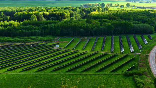 Aerial view of solar panels arranged in rows across a lush green field bordered by dense forest and a road in Latvia. Aerial view of solar panels arranged in rows across a lush green field bordered by dense forest and a road in Latvia.