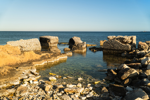 View of weathered concrete blocks and rocky shoreline along the clear Mediterranean sea under a bright blue sky in Marsaskala Southern Region Malta. View of weathered concrete blocks and rocky shoreline along the clear Mediterranean sea under a bright blue sky in Marsaskala Southern Region Malta.