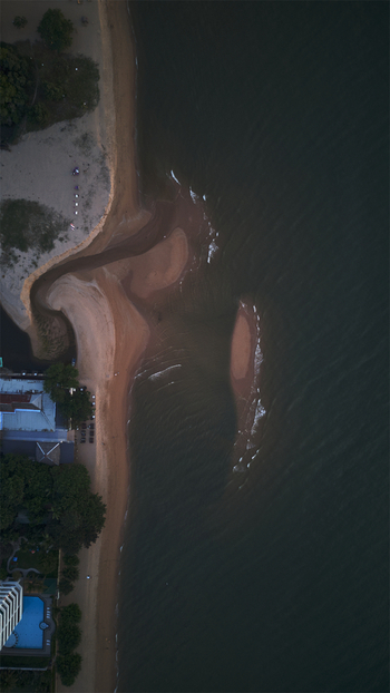 Aerial view of Jomtien Beach featuring the sandy shoreline where a small river mouth meets the dark ocean water near coastal buildings and trees in Chon Buri Thailand.