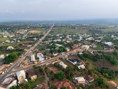 Aerial view of a long road splitting the landscape with buildings clustered along the roadside and lush green vegetation covering the distant hills Elu Abia Nigeria. Aerial view of a long road splitting the landscape with buildings clustered along the roadside and lush green vegetation covering the distant hills Elu Abia Nigeria.