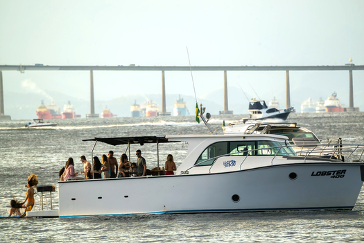 Rio de Janeiro Brazil 05 June 2026 View of a white motorboat with people on deck cruising in Guanabara Bay with the Rio Niteroi Bridge and cargo ships in the background.