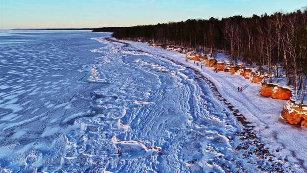 Aerial view of a frozen sea and ice covered coastline with sandy cliffs pine trees and people walking along the shore in Latvia. Aerial view of a frozen sea and ice covered coastline with sandy cliffs pine trees and people walking along the shore in Latvia.