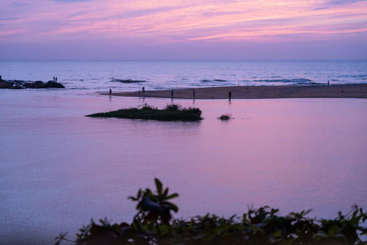View of a purple sunset over a coastal lagoon with silhouettes of people on the beach and twilight sky reflecting on the water in Galle Southern Province Sri Lanka.