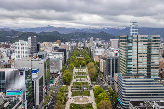 Aerial view of Odori Park stretching through the city center flanked by modern skyscrapers lush green trees and distant mountains under a cloudy sky in Sapporo Hokkaido Japan. Aerial view of Odori Park stretching through the city center flanked by modern skyscrapers lush green trees and distant mountains under a cloudy sky in Sapporo Hokkaido Japan.