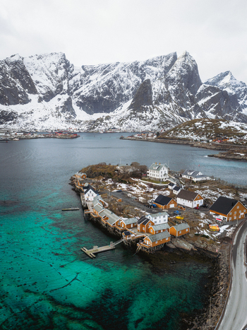 Aerial view of vibrant turquoise waters meet the rustic charm of wooden houses nestled at the foot of snow dusted mountains Sakrisøya Nordland Norway. Aerial view of vibrant turquoise waters meet the rustic charm of wooden houses nestled at the foot of snow dusted mountains Sakrisøya Nordland Norway.