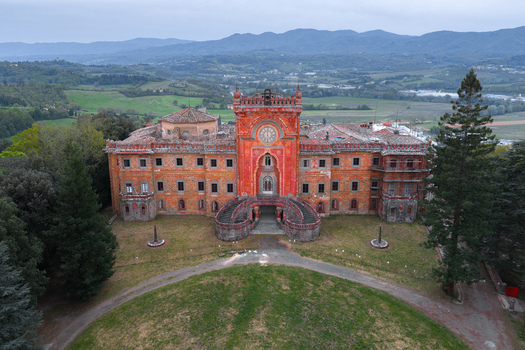 Aerial view of the historic Castello di Sammezzano with its red facade and clock tower surrounded by lush green trees and rolling hills in Leccio Toscana Italy. Aerial view of the historic Castello di Sammezzano with its red facade and clock tower surrounded by lush green trees and rolling hills in Leccio Toscana Italy.