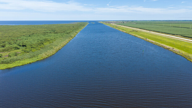 Aerial view of the canal leading into Lake Okeechobee flanked by lush green vegetation and agricultural fields under a clear blue sky in Pahokee Florida United States. Aerial view of the canal leading into Lake Okeechobee flanked by lush green vegetation and agricultural fields under a clear blue sky in Pahokee Florida United States.