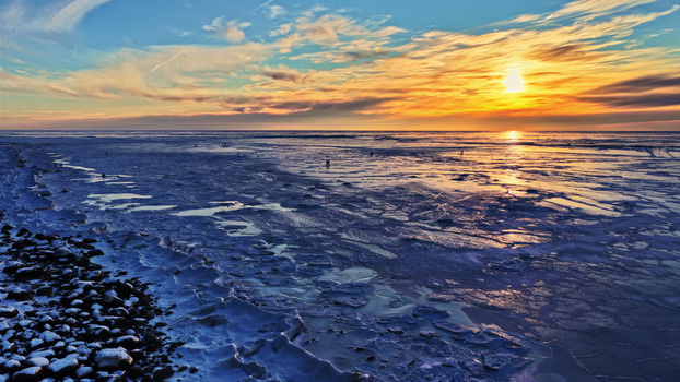 Aerial view of a frozen icy coastline at sunset with vibrant orange and blue sky reflecting on the ice and dark pebbles along the shore in Latvia. Aerial view of a frozen icy coastline at sunset with vibrant orange and blue sky reflecting on the ice and dark pebbles along the shore in Latvia.