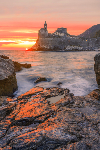 Aerial view of Chiesa di San Pietro perched on a rocky cliff during a vibrant sunset with orange skies and waves crashing against the shore in Portovenere Liguria Italy. Aerial view of Chiesa di San Pietro perched on a rocky cliff during a vibrant sunset with orange skies and waves crashing against the shore in Portovenere Liguria Italy.