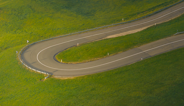 Aerial view of a dramatic hairpin mountain road crossing green alpine meadows in Italy.