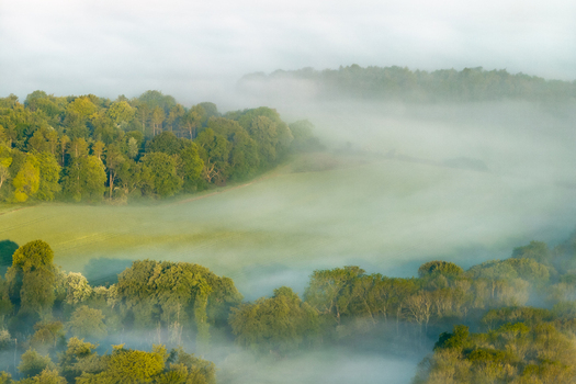 Aerial view of mist rolling over green fields and dense woodland in the rolling hills of the countryside near Nuffield Oxfordshire United Kingdom. Aerial view of mist rolling over green fields and dense woodland in the rolling hills of the countryside near Nuffield Oxfordshire United Kingdom.