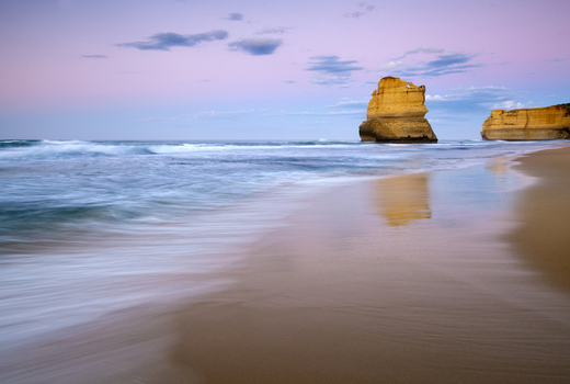 View of the Twelve Apostles limestone stacks rising from the ocean waves along a sandy beach under a purple twilight sky Australia. View of the Twelve Apostles limestone stacks rising from the ocean waves along a sandy beach under a purple twilight sky Australia.