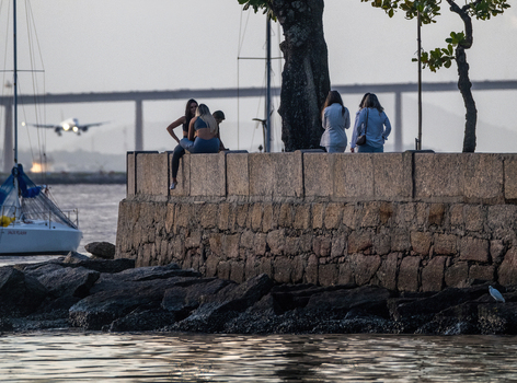 Rio de Janeiro Brazil 05 June 2026 View of people sitting on the Mureta da Urca stone wall with an airplane landing and the Rio Niteroi Bridge in the background.