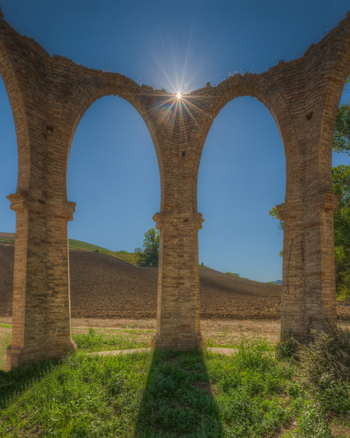 View of ancient stone arches with a sunburst shining through the structure set against a rural landscape with plowed fields and a clear blue sky in Ponzano di Fermo Marche Italy. View of ancient stone arches with a sunburst shining through the structure set against a rural landscape with plowed fields and a clear blue sky in Ponzano di Fermo Marche Italy.