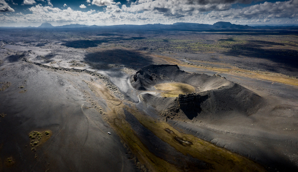 Aerial view of a volcanic crater surrounded by dark ash fields and rugged terrain under a cloudy sky in Iceland.