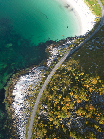 Aerial view of the winding coastal road along the turquoise waters and white sand of Vik Strand with rocky shores and autumn trees in Leknes Nordland Norway. Aerial view of the winding coastal road along the turquoise waters and white sand of Vik Strand with rocky shores and autumn trees in Leknes Nordland Norway.
