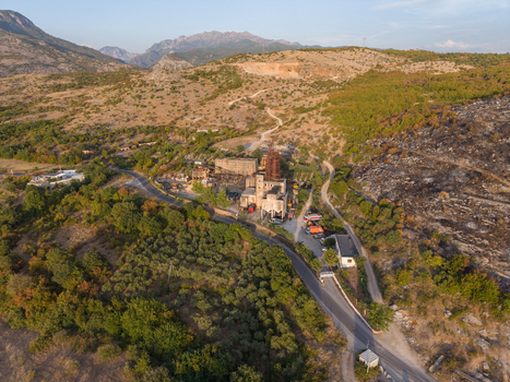 Aerial view of an industrial facility nestled in a mountainous landscape with olive groves a winding road and signs of recent vegetation fire on the hillside in Boks Qarku i Shkodres Albania.