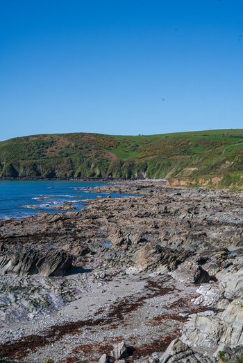 View of the rocky coastline at Portnadler Bay along the South West Coastal Path with green hills and blue sky in Looe Cornwall United Kingdom. View of the rocky coastline at Portnadler Bay along the South West Coastal Path with green hills and blue sky in Looe Cornwall United Kingdom.