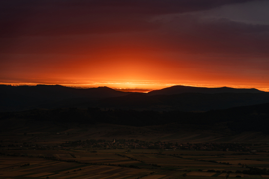 View of a vibrant sunset casting a warm orange glow over the rolling hills and agricultural fields surrounding the village of Mereni Covasna County Romania. View of a vibrant sunset casting a warm orange glow over the rolling hills and agricultural fields surrounding the village of Mereni Covasna County Romania.