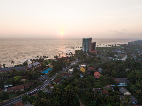 Aerial view of the coastal town featuring lush palm trees residential houses and a tall modern building along the ocean shore during a hazy sunset Galle Southern Province Sri Lanka. Aerial view of the coastal town featuring lush palm trees residential houses and a tall modern building along the ocean shore during a hazy sunset Galle Southern Province Sri Lanka.