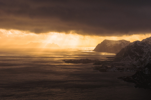 Aerial view of sun rays pierce through dark clouds illuminating the rugged coastline and tranquil waters of Reinebringen Nordland Norway.