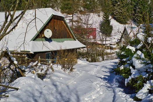 View of a snow covered path leading past a rustic wooden cabin under a bright sky nestled among snow dusted trees creating a winter wonderland Moce Banska Bystrica Region Slovakia.