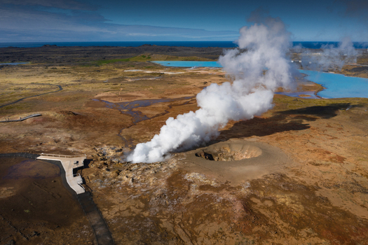 Aerial view of a geothermal area with a massive steam plume rising from a volcanic vent near blue pools and a wooden walkway in Iceland.