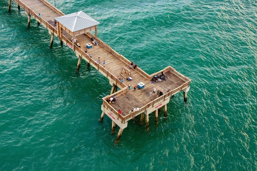 Aerial view of the T shaped Deerfield Beach International Fishing Pier extending over turquoise ocean water with people fishing and walking in Deerfield Beach Florida United States.