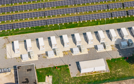 Aerial view of battery energy storage systems integrated with a large solar farm featuring rows of photovoltaic panels and white storage containers on a gravel site in Italy.