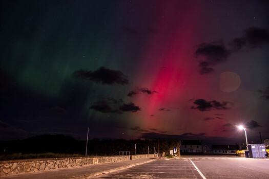 View of the Aurora Borealis illuminating the night sky with vibrant green and red lights above a coastal parking area and stone wall in Portnoo County Donegal Ireland. View of the Aurora Borealis illuminating the night sky with vibrant green and red lights above a coastal parking area and stone wall in Portnoo County Donegal Ireland.