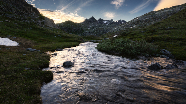 View of a mountain stream flowing through a rocky valley with green slopes and jagged peaks under a blue sky in the Aosta Valley Italy. View of a mountain stream flowing through a rocky valley with green slopes and jagged peaks under a blue sky in the Aosta Valley Italy.