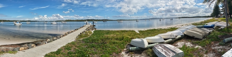 Aerial view of boats resting on the shore contrasting with the blue water and sky and a concrete pier stretching into the bay Port Stephens Australia.