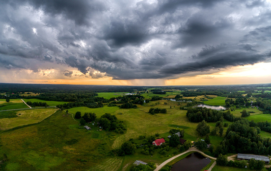 Aerial view of a rural landscape in Latvia featuring green fields scattered trees small ponds and houses under a dramatic storm sky with rain showers at sunset Latvia. Aerial view of a rural landscape in Latvia featuring green fields scattered trees small ponds and houses under a dramatic storm sky with rain showers at sunset Latvia.