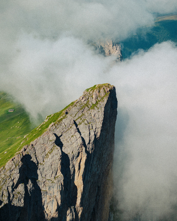 Aerial view of the sharp mountain peak of Seceda rising above thick white clouds with green alpine meadows in Ortisei Trentino Alto Adige Italy.