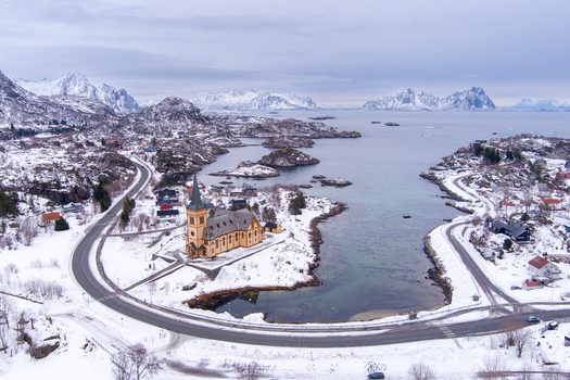 Aerial view of Vagan Church in Lofoten surrounded by snow covered mountains icy waters and winding roads in the winter landscape of Nordland Norway.