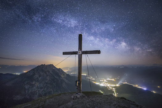 View of the summit cross on Stoderzinken mountain under the Milky Way and starry night sky overlooking the valley lights Liezen Steiermark Austria. View of the summit cross on Stoderzinken mountain under the Milky Way and starry night sky overlooking the valley lights Liezen Steiermark Austria.