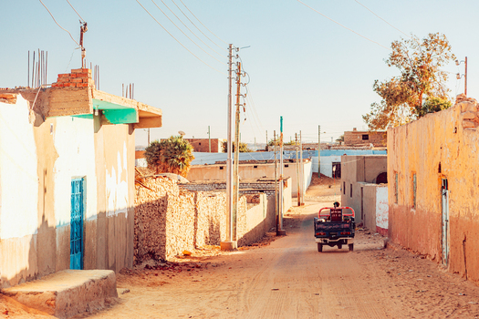 View of a dusty village street with traditional earth toned houses and a small vehicle driving under a clear sky Aswan Aswan Governorate Egypt. View of a dusty village street with traditional earth toned houses and a small vehicle driving under a clear sky Aswan Aswan Governorate Egypt.