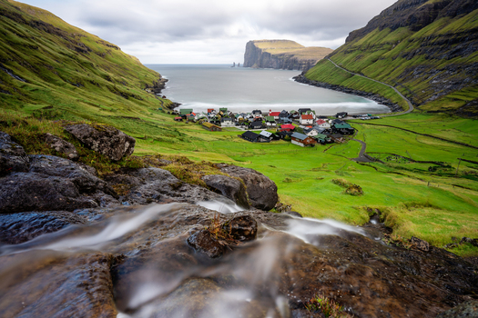 View of the village of Tjornuvik nestled between lush green hills and a rocky bay featuring a cascading waterfall in the foreground Faroe Islands.