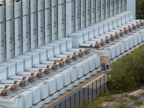 Aerial view of the exterior cooling infrastructure of a data center featuring rows of industrial ventilation units and metal piping along a perimeter fence in the United Kingdom.