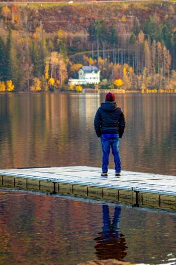 View of a man standing on a wooden pier looking out over a calm lake with autumn trees and a house in the background Breitnau Baden Wurttemberg Germany. View of a man standing on a wooden pier looking out over a calm lake with autumn trees and a house in the background Breitnau Baden Wurttemberg Germany.