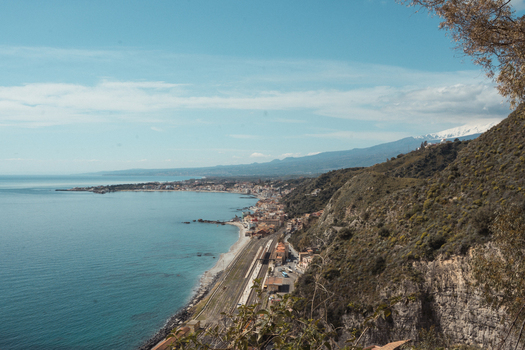 View of the coastline meeting the sea the mountain ranges and the town nestled between under a bright sky Taormina Sicily Italy.