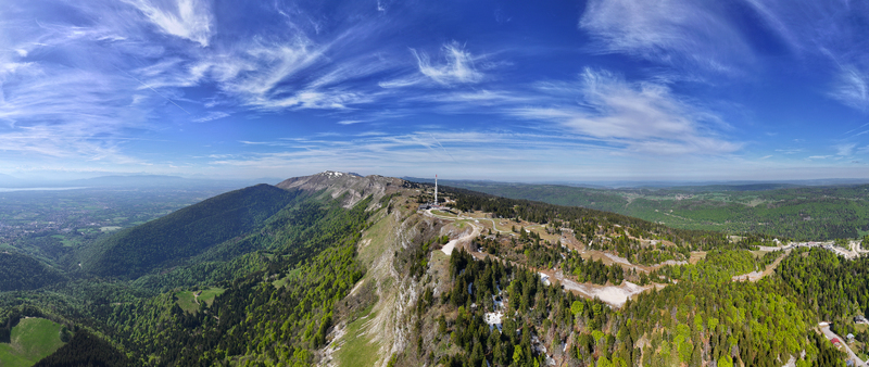 Aerial view of Col de la Faucille mountain pass featuring a telecommunications tower and lush green forests under a blue sky with wispy clouds in Gex Auvergne Rhone Alpes France. Aerial view of Col de la Faucille mountain pass featuring a telecommunications tower and lush green forests under a blue sky with wispy clouds in Gex Auvergne Rhone Alpes France.
