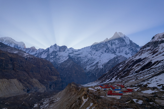 Aerial view of the snow capped Machapuchare peak in the Himalayas with sun rays breaking over the summit and a base camp with red roofed buildings in Nepal.