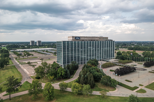 Dearborn United States 04 August 2024 Aerial view of the Ford World Headquarters building surrounded by green lawns parking lots and flags under a cloudy sky.