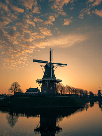 Aerial view of a traditional windmill silhouetted against a golden sunset sky with scattered clouds and a clear reflection on the calm water in Dokkum Friesland Netherlands. Aerial view of a traditional windmill silhouetted against a golden sunset sky with scattered clouds and a clear reflection on the calm water in Dokkum Friesland Netherlands.