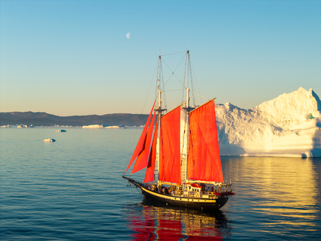 Aerial view of a schooner with vibrant red sails navigating through calm Arctic waters surrounded by massive icebergs under a clear blue sky in Greenland. Aerial view of a schooner with vibrant red sails navigating through calm Arctic waters surrounded by massive icebergs under a clear blue sky in Greenland.