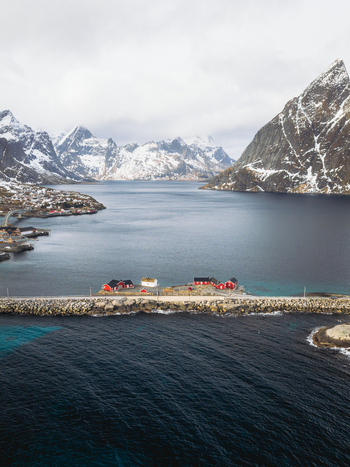 Aerial view of red cabins stand out against the deep blue waters with snow capped mountains rising majestically in the background Sakrisøya Nordland Norway. Aerial view of red cabins stand out against the deep blue waters with snow capped mountains rising majestically in the background Sakrisøya Nordland Norway.